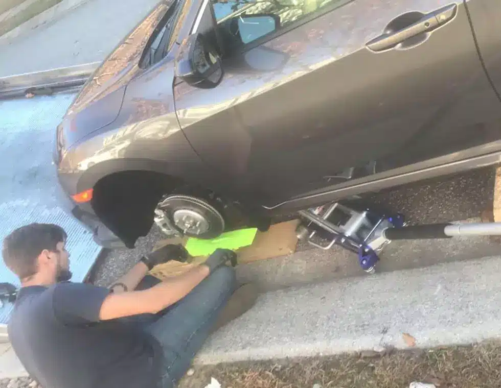 A man sitting beside a car, changing its tire. Car tire change service being performed by a person near a parked vehicle.