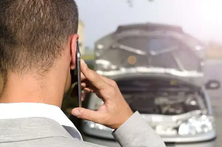 A person stands beside a car, speaking on the phone to a towing service provider for assistance.