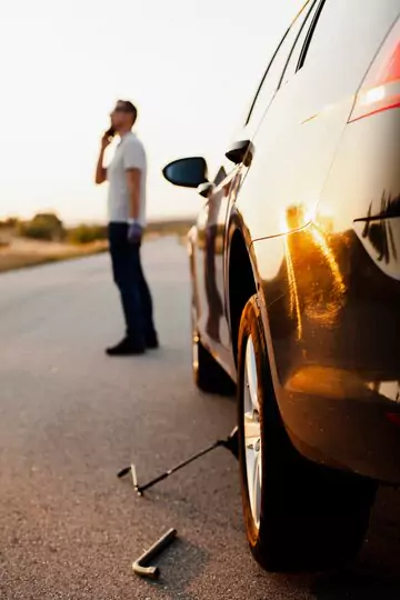 A man standing on the roadside next to a car, holding a mobile phone and calling a towing company for roadside assistance.